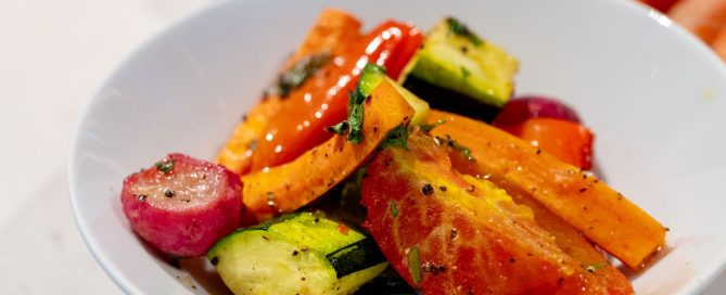 Een foto van een wit schaaltje op een werkblad van een keuken. In het schaaltje bevinden zich gegrilde groenten met wat groene versiering, in de achtergrond zien we de rauwe groenten onscherp liggen.