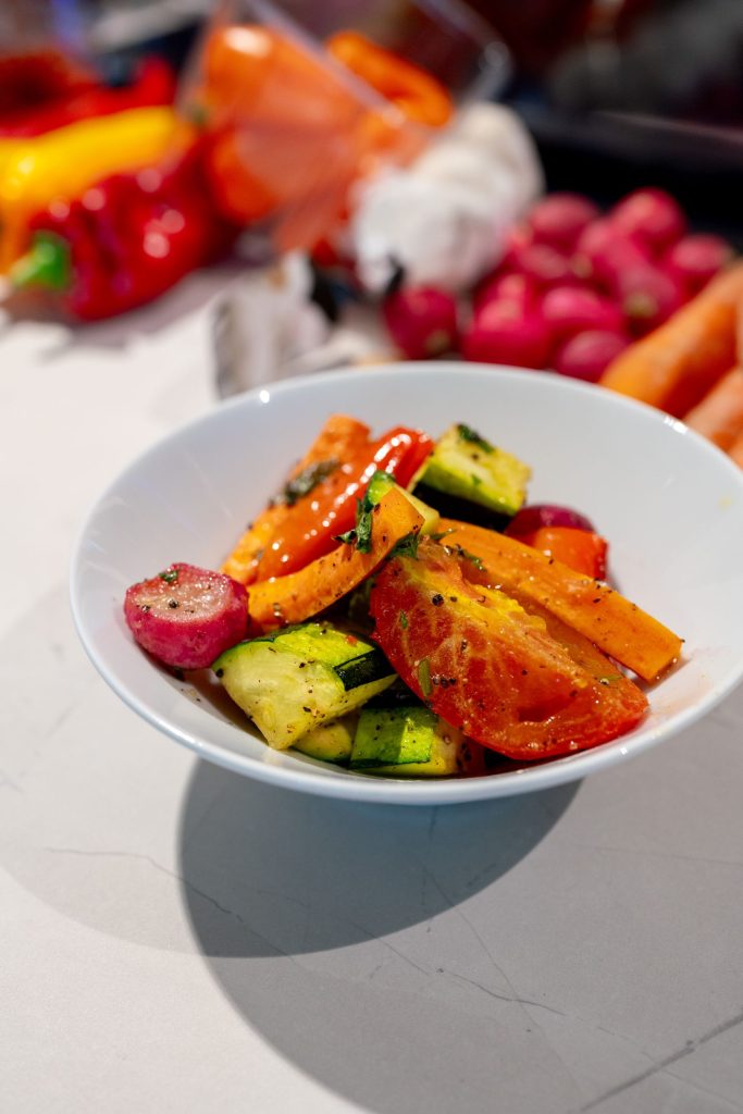 Een foto van een wit schaaltje op een werkblad van een keuken. In het schaaltje bevinden zich gegrilde groenten met wat groene versiering, in de achtergrond zien we de rauwe groenten onscherp liggen.